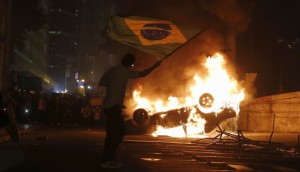 A demonstrator waves a Brazilian flag by a burning a car in downtown Rio de Janeiro